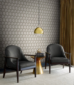 Two black armchairs sit beside a small gold table with a decorative object, under a hanging gold lamp. The background features York Wallcoverings’ Coco Bloom Wallpaper and gold curtains.