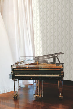 A transparent acrylic grand piano with gold accents sits on a polished wooden floor near white curtains and York Wallcoverings’ Metropolis Wallpaper.