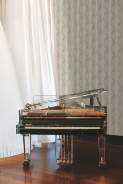 A transparent grand piano with gold accents sits on a polished wooden floor, near white curtains and Metropolis Wallpaper by York Wallcoverings.