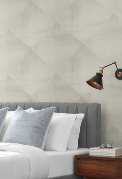 In Clay Wallpaper with a subtle, large-scale diamond pattern behind a bed with a gray tufted headboard and a black wall-mounted lamp.