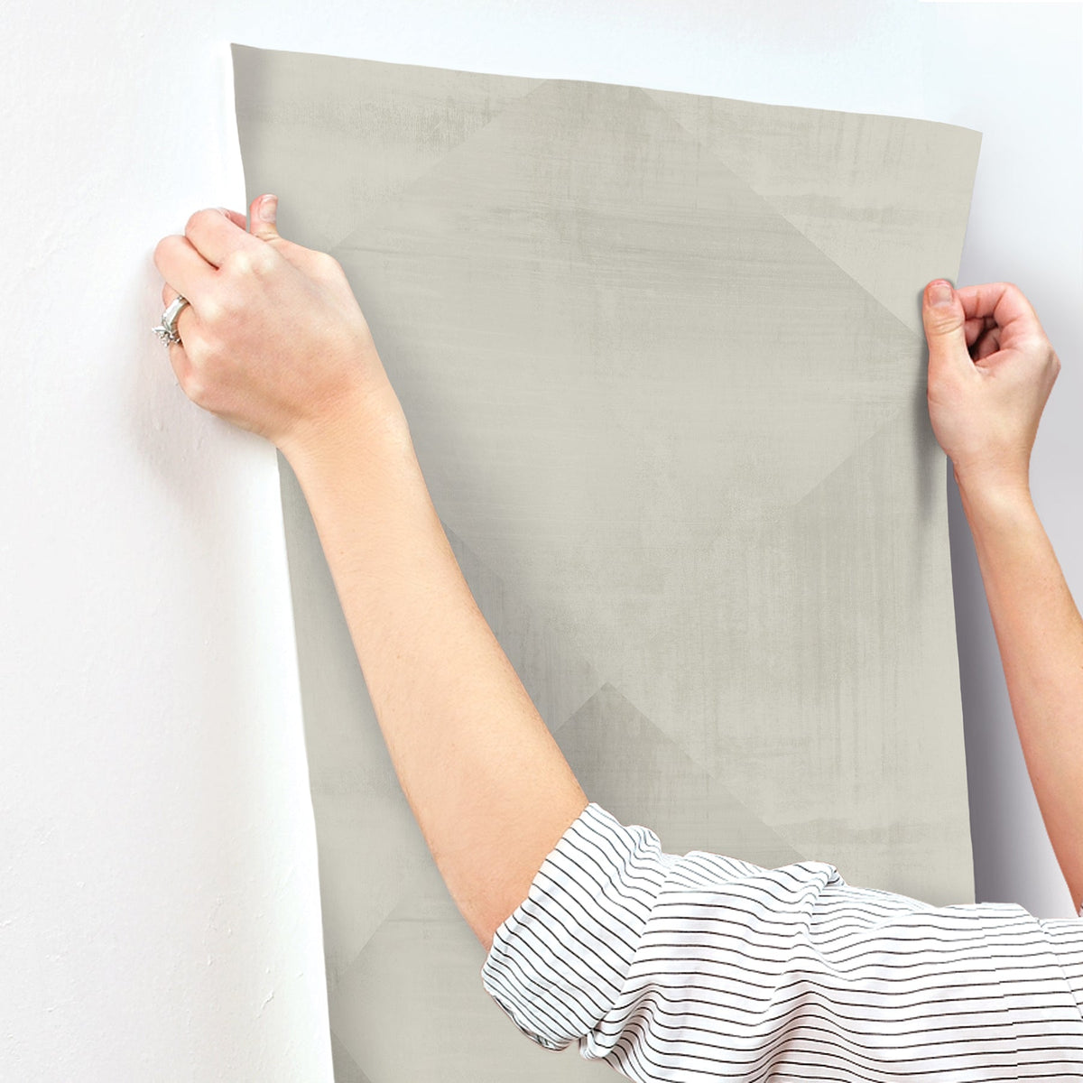 Hands holding a sheet of light gray In Clay Wallpaper with a subtle geometric pattern against a wall.