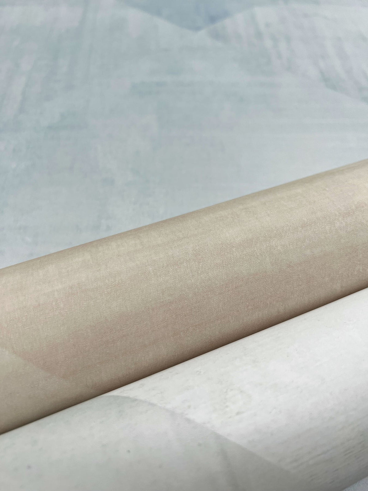 A roll of beige In Clay Wallpaper shown against a background of blue and white patterned wallpaper.