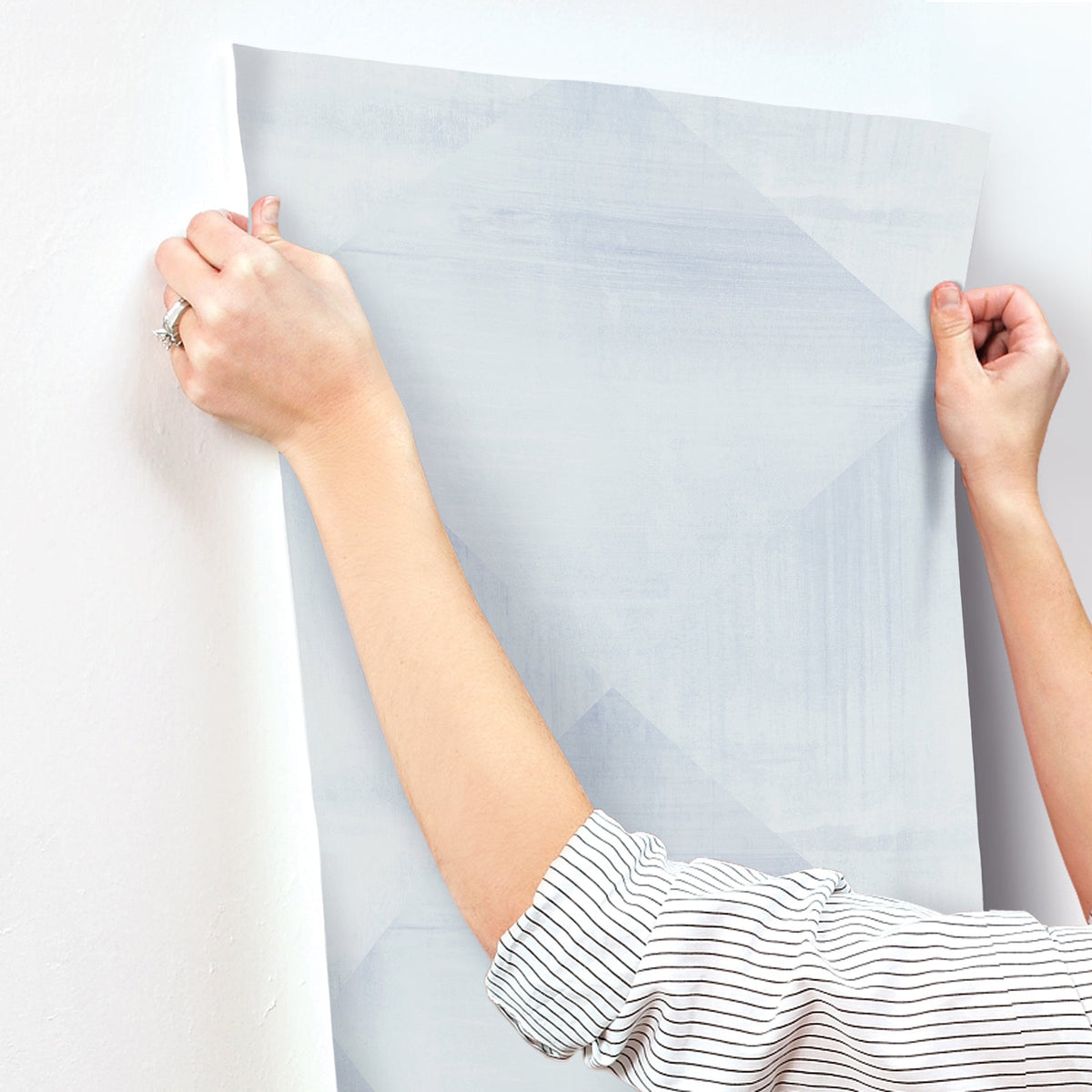 Hands applying a sheet of light blue geometric-patterned In Clay Wallpaper by York Wallcoverings to a wall.