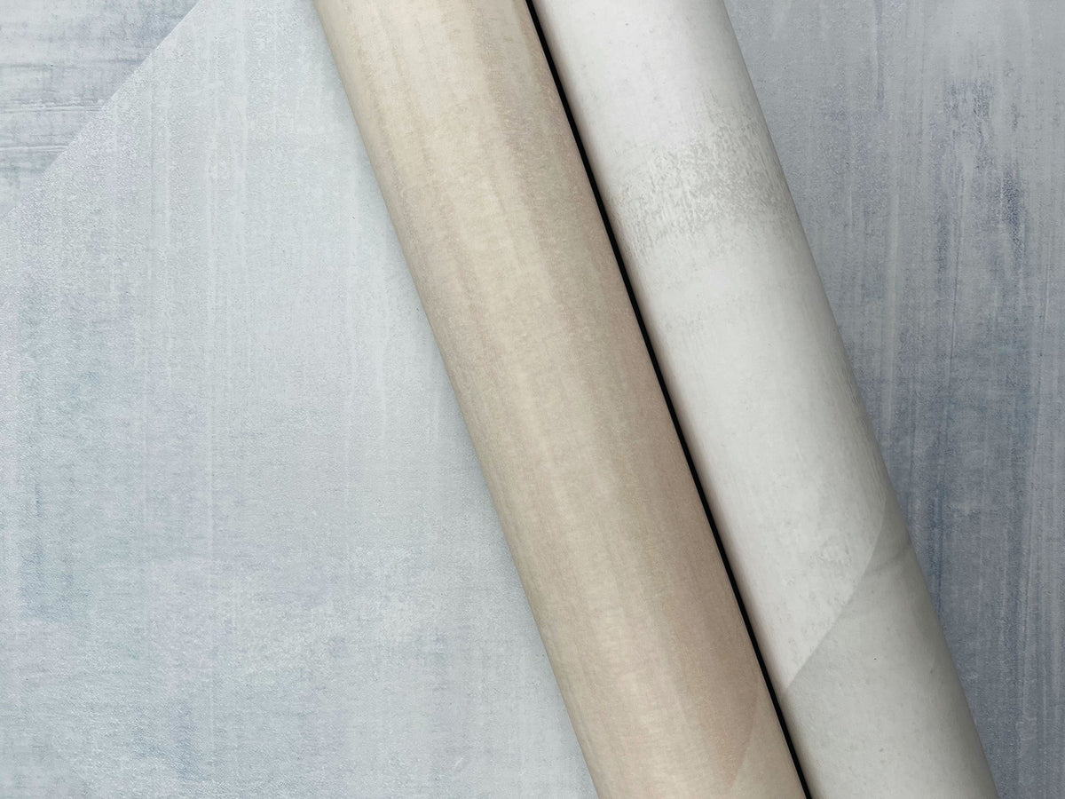 Two rolls of In Clay Wallpaper with a mottled, textured appearance, one in beige and the other in white, resting on a blue-grey background.