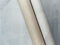 Two rolls of In Clay Wallpaper with a mottled, textured appearance, one in beige and the other in white, resting on a blue-grey background.