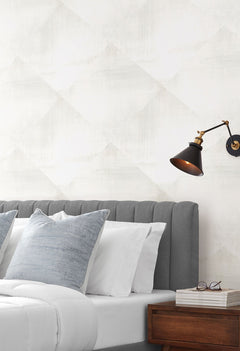 In Clay Wallpaper with a subtle, light gray diamond pattern behind a bed with gray upholstery and a black wall sconce.