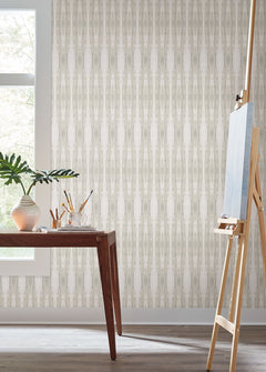 A wooden table adorned with art supplies and a plant sits by a window; an empty easel stands nearby against a backdrop of York Wallcoverings’ Escalante Wallpaper.