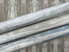 Three rolls of Escalante Wallpaper by York Wallcoverings, in grey, cream, and blue hues, are stacked diagonally against a coordinated patterned backdrop. Each roll displays a soft abstract design with brushstroke-like accents.