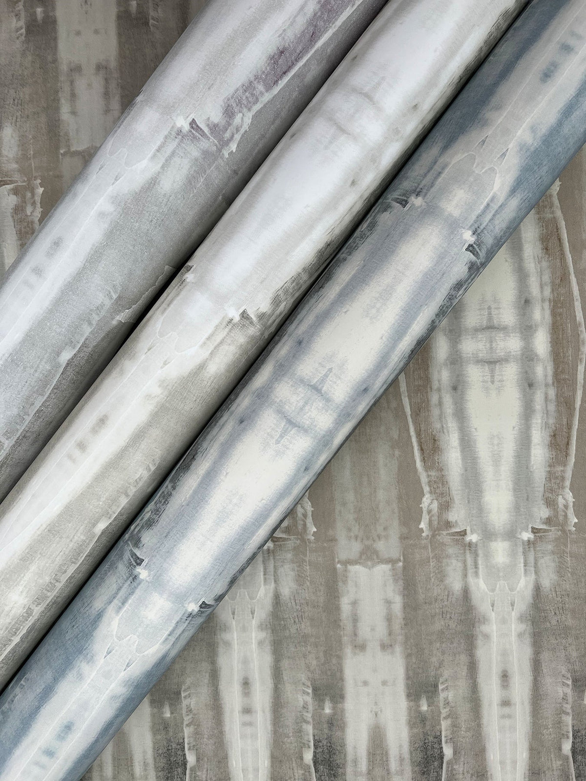 Three rolls of Escalante Wallpaper with a mottled, grey and white watercolor-style pattern resting against a matching wallpaper background.