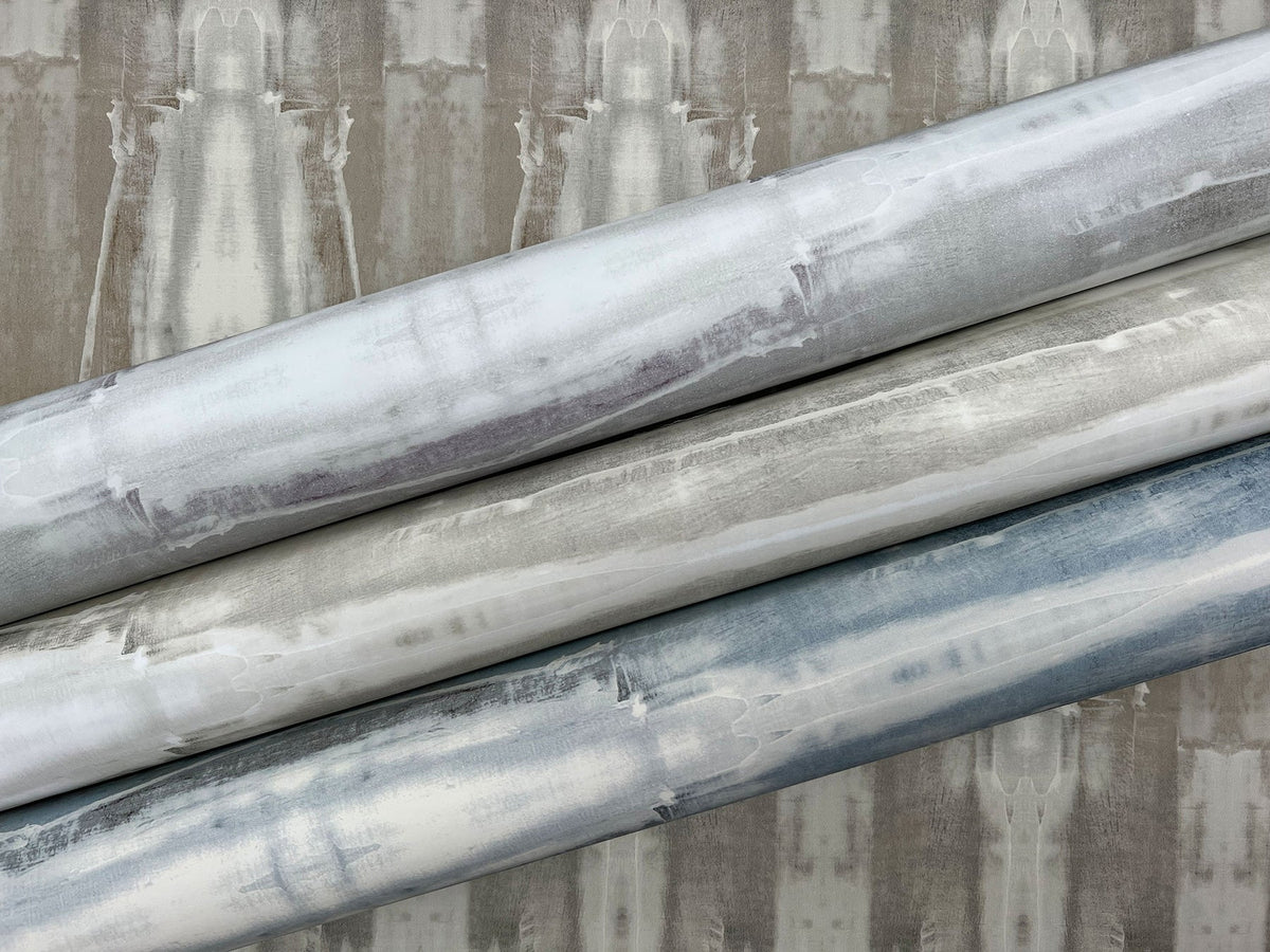 Three rolls of Escalante Wallpaper by York Wallcoverings featuring a blurred, vertical abstract pattern in shades of grey, blue, and white, displayed against a matching background.
