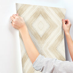 Hands applying a sheet of Ballad Wallpaper with a beige and white diamond-patterned brushstroke design to a white wall.