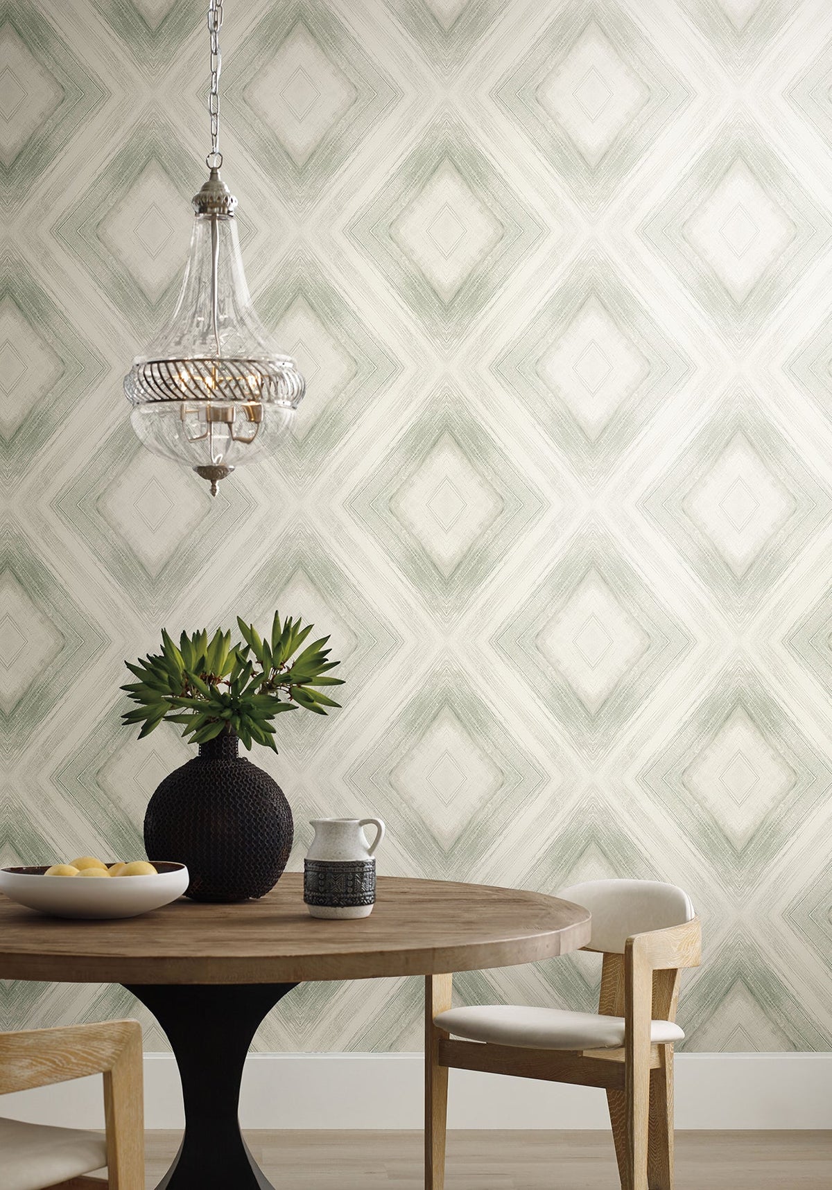 Ballad Wallpaper featuring a repeating sage green diamond pattern behind a round wooden table, a dark vase with greenery, and a glass pendant light.