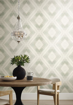 Ballad Wallpaper featuring a repeating sage green diamond pattern behind a round wooden table, a dark vase with greenery, and a glass pendant light.
