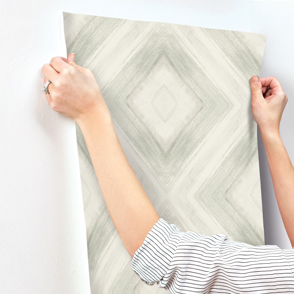 Hands holding up a sheet of Ballad Wallpaper, featuring a gray and white diamond-shaped brushstroke pattern, against a wall.