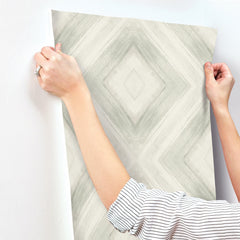 Hands holding up a sheet of Ballad Wallpaper, featuring a gray and white diamond-shaped brushstroke pattern, against a wall.