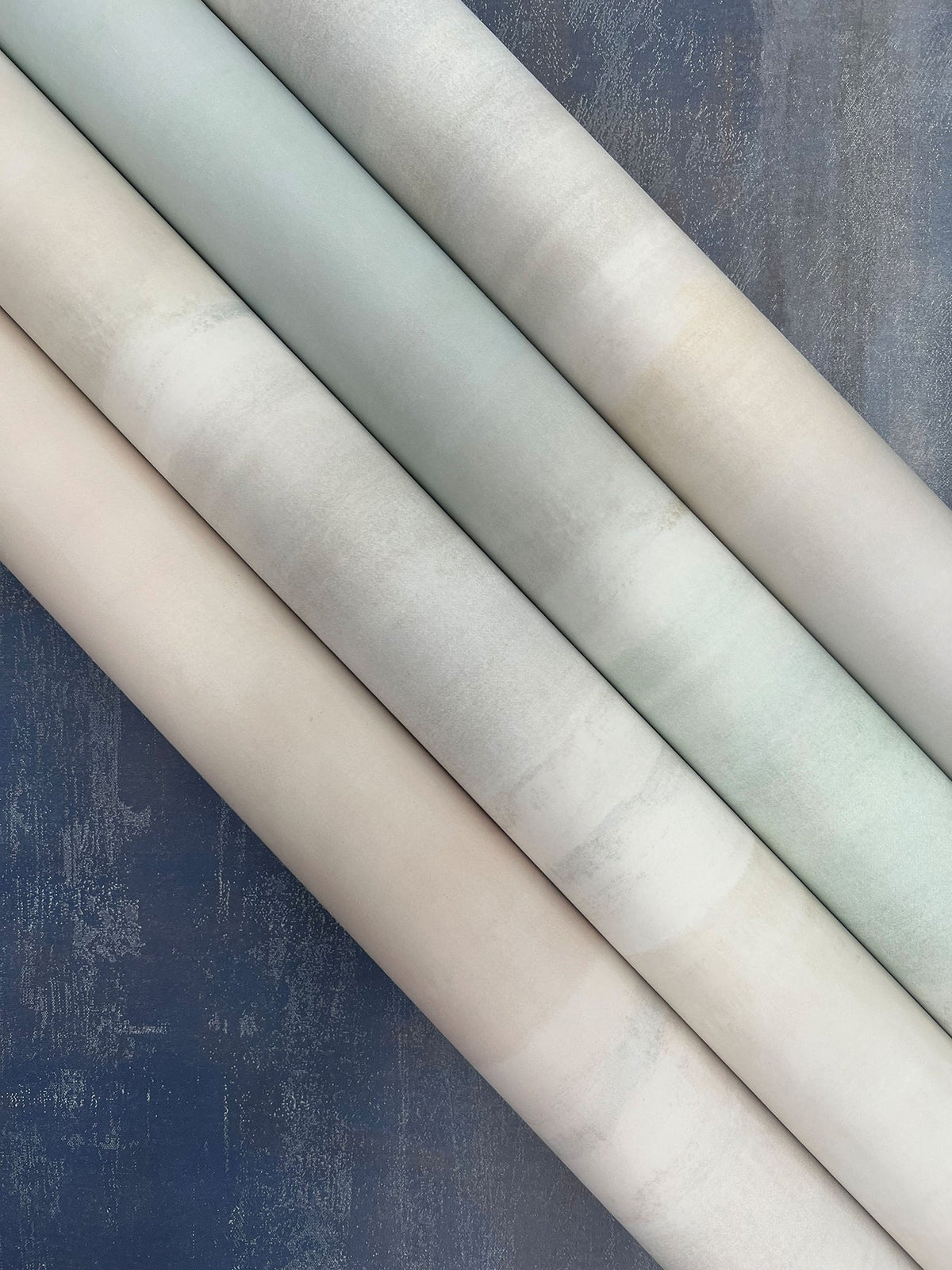 Four rolls of Serene Reflection Wallpaper in shades of cream, grey, and pale blue arranged diagonally across a dark blue textured background.