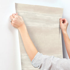 Hands holding a sheet of light grey, horizontally-streaked York Wallcoverings Grey Stone Wallpaper against a white wall.