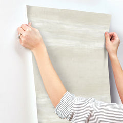 Hands holding a sheet of light grey, textured Grey Stone Wallpaper against a white wall.