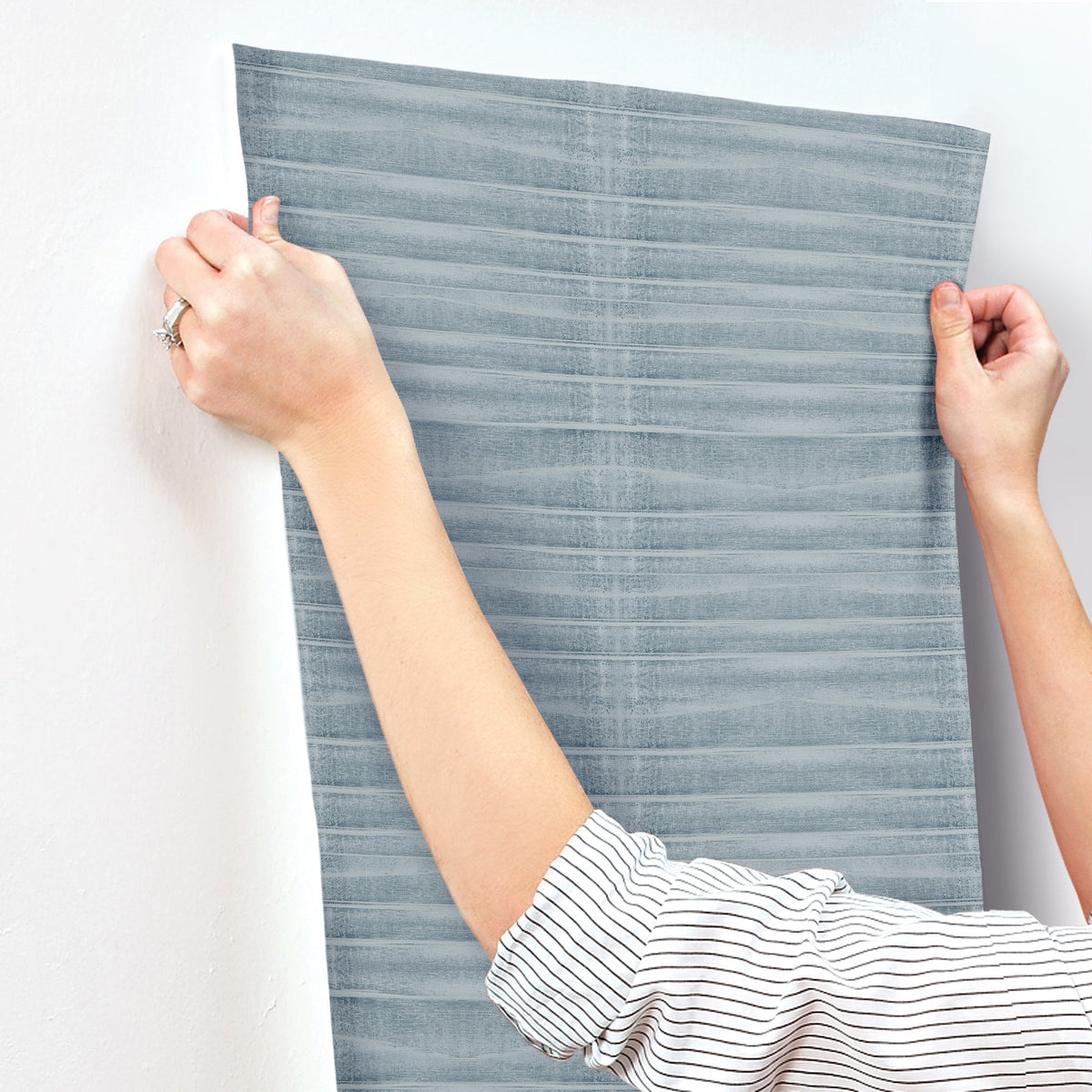 Hands apply a strip of Grey Stone Wallpaper, featuring a horizontal, textured pattern, to a white wall.