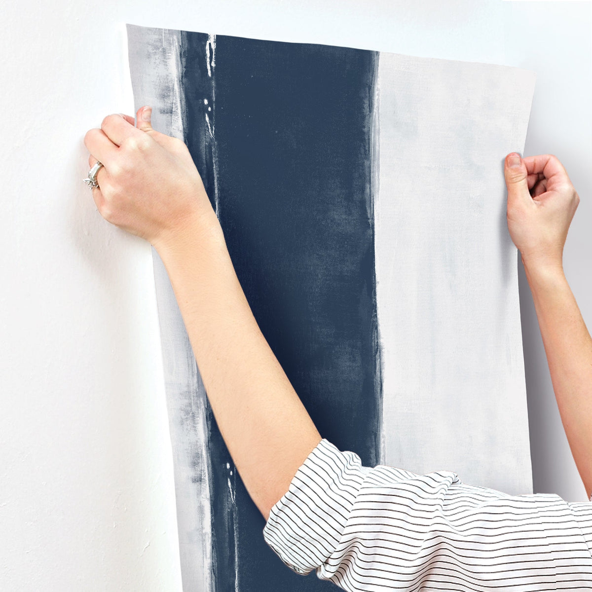 Hands holding a strip of Ink Wash Wallpaper by York Wallcoverings, featuring a wide navy blue vertical stripe on a light gray background.