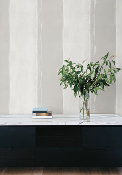 Ink Wash Wallpaper by York Wallcoverings features vertical stripes in shades of gray and white, positioned behind a black sideboard holding a glass vase of greenery and a stack of books.