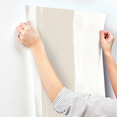 Hands applying a strip of Ink Wash Wallpaper by York Wallcoverings, featuring a wide, vertical beige stripe against a white background, to a wall.