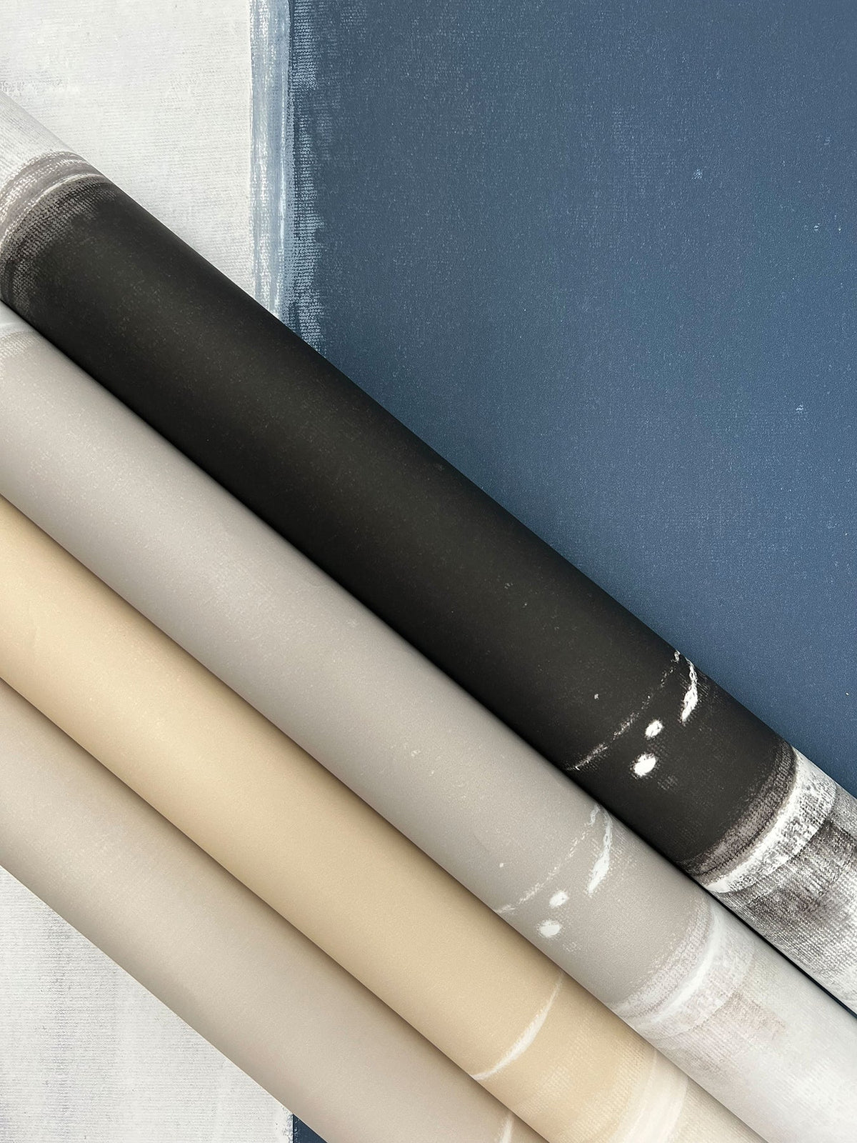 Four diagonal rolls of Ink Wash Wallpaper in gradient shades of black, gray, beige, and white, resting against a painted blue and white surface.