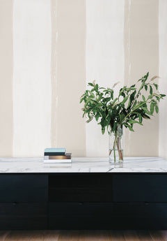 Ink Wash Wallpaper features wide, vertical brushstroke stripes in alternating shades of beige and off-white behind a black console table with a marble top, a stack of books, and a glass vase of greenery.