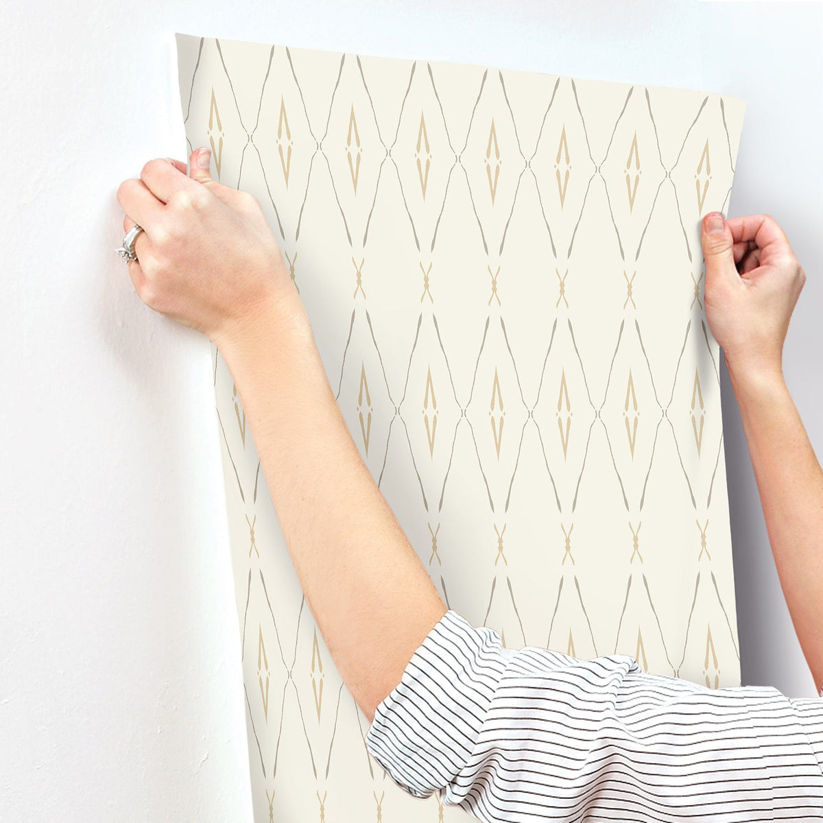 Hands applying cream-colored Artifact Silhouettes Wallpaper with a tan geometric diamond pattern to a white wall.