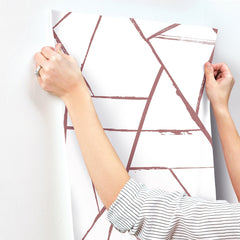 Hands applying a sheet of York Wallcoverings Intersect Wallpaper with a geometric pattern of muted pink intersecting lines to a wall.