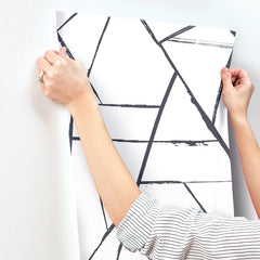Hands applying a sheet of Intersect Wallpaper, featuring a white background with a black geometric pattern of intersecting lines, to a wall.
