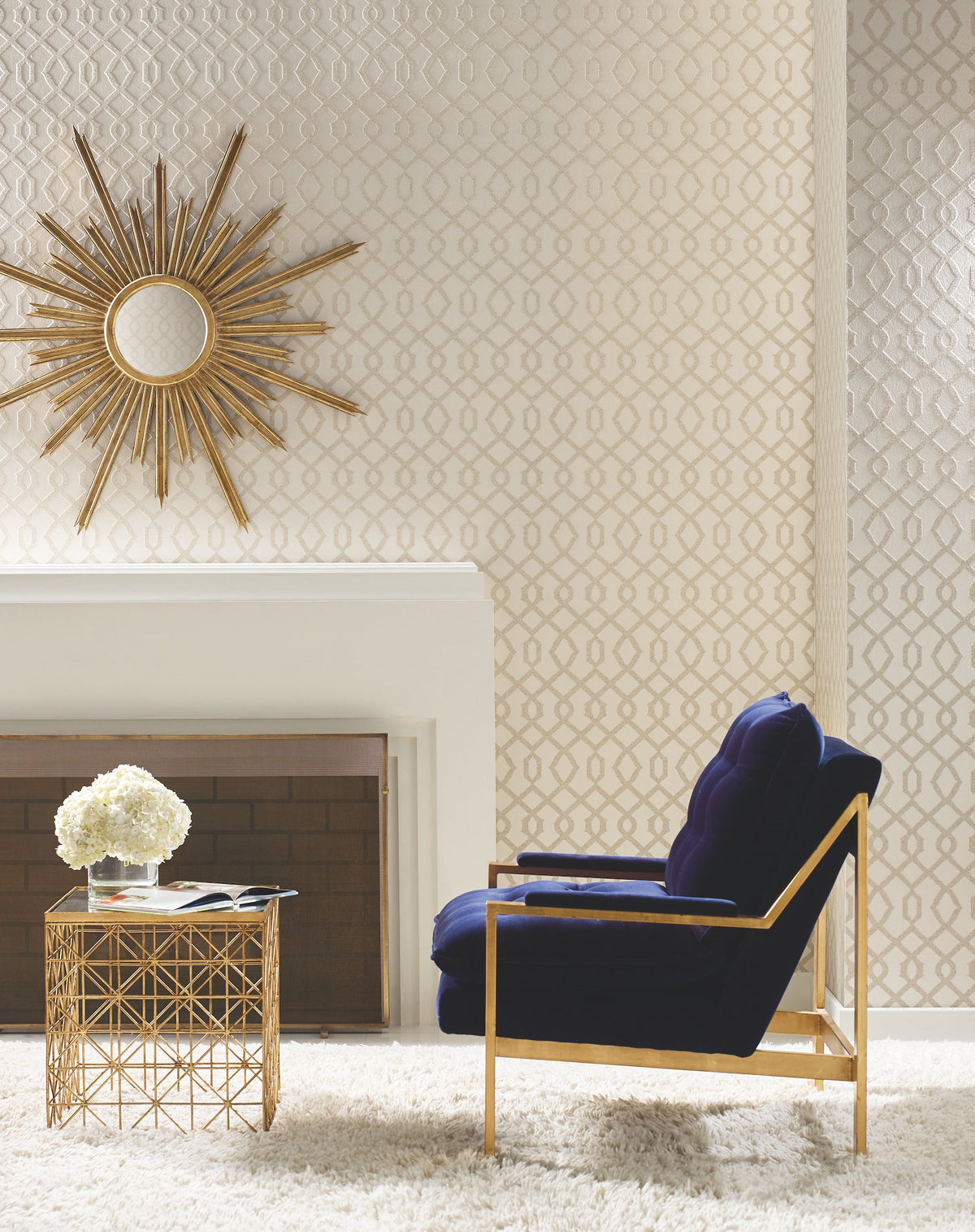 Luscious Wallpaper by York Wallcoverings features a repeating geometric diamond pattern on a wall next to a fireplace, a sunburst mirror, and a dark blue armchair.