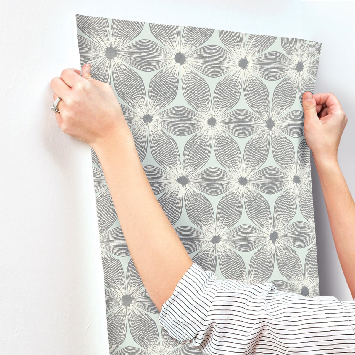 Hands apply Candice Olson Everlasting Wallpaper by York Wallcoverings, featuring a light green and grey floral pattern, to a white wall.