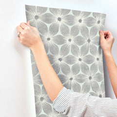 Hands apply Candice Olson Everlasting Wallpaper by York Wallcoverings, featuring a light green and grey floral pattern, to a white wall.