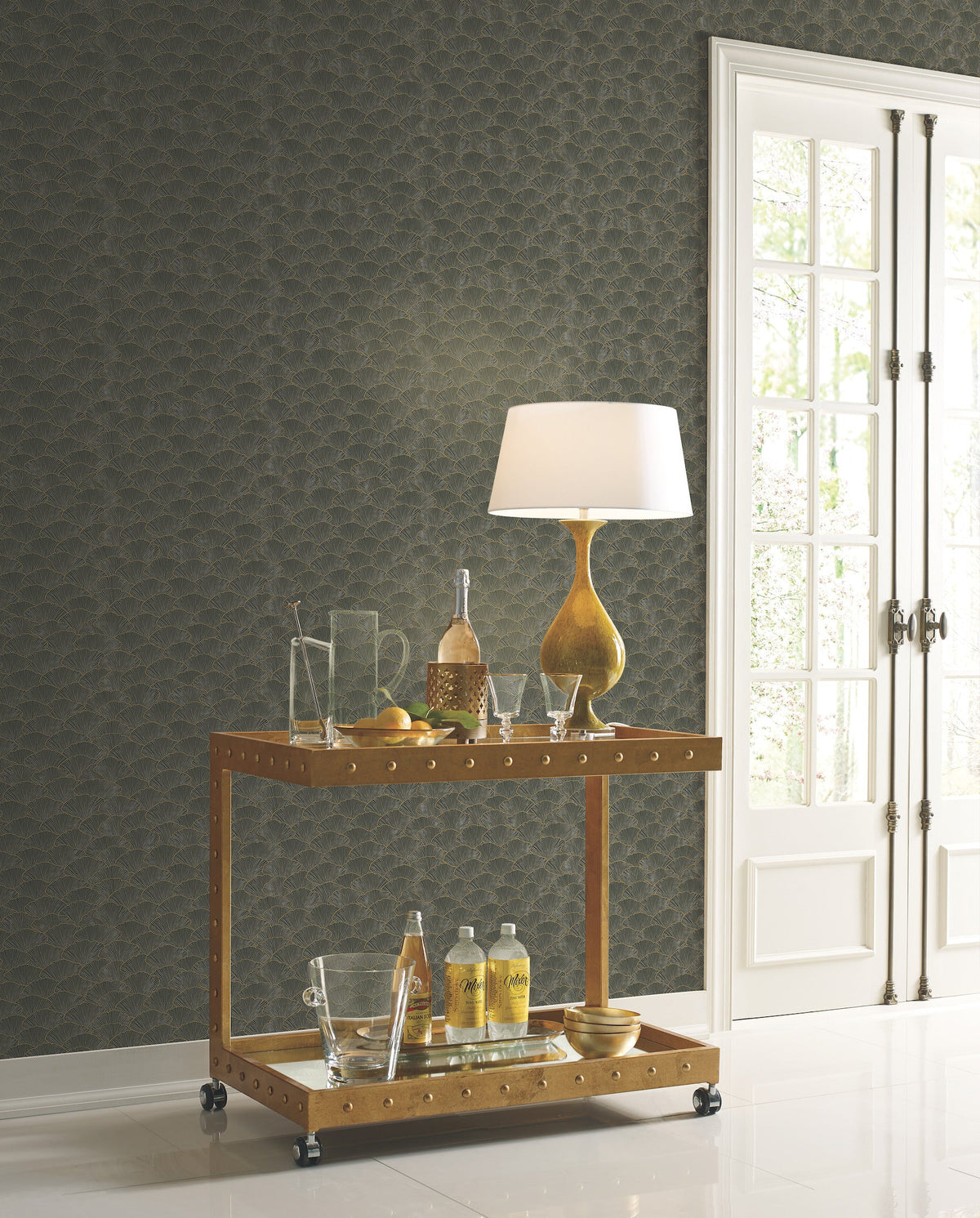 A bar cart topped with a lamp and glassware stands in front of a dark gray wall featuring Candice Olson Luminous Gingko Wallpaper with a gold fan pattern.