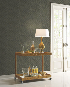 A bar cart topped with a lamp and glassware stands in front of a dark gray wall featuring Candice Olson Luminous Gingko Wallpaper with a gold fan pattern.