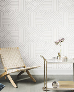 Candice Olson Diverging Diamonds Wallpaper features a geometric pattern of overlapping thin, light-colored diamond lines on a white background, displayed behind a wooden woven chair and a rolling metal cart.