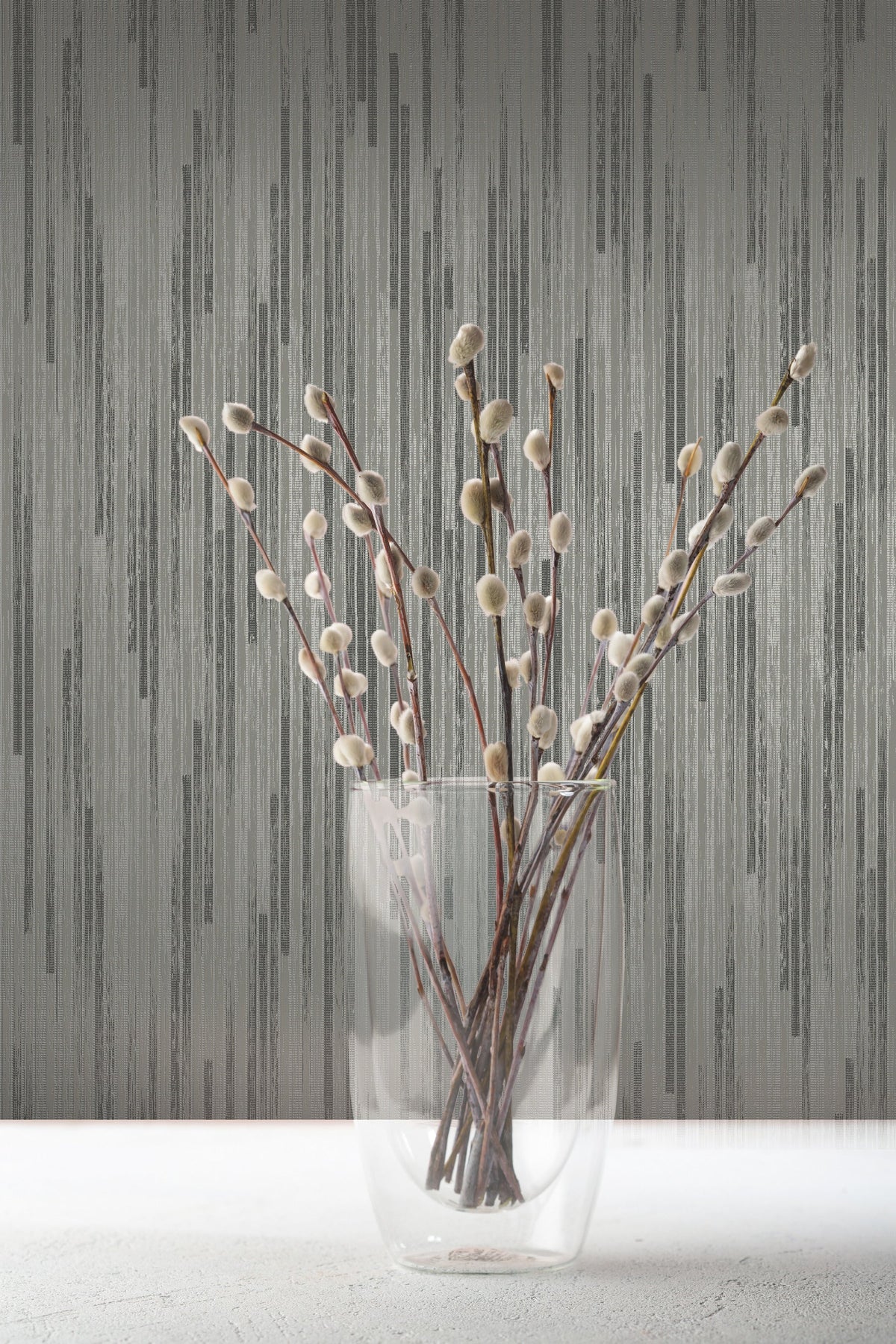 A clear glass vase holding pussy willow branches in front of gray Candice Olson Bargello Wallpaper with a vertical textured pattern.