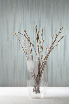 Clear glass vase containing pussy willow branches in front of blue textured Candice Olson Bargello Wallpaper by York Wallcoverings with vertical stripes.