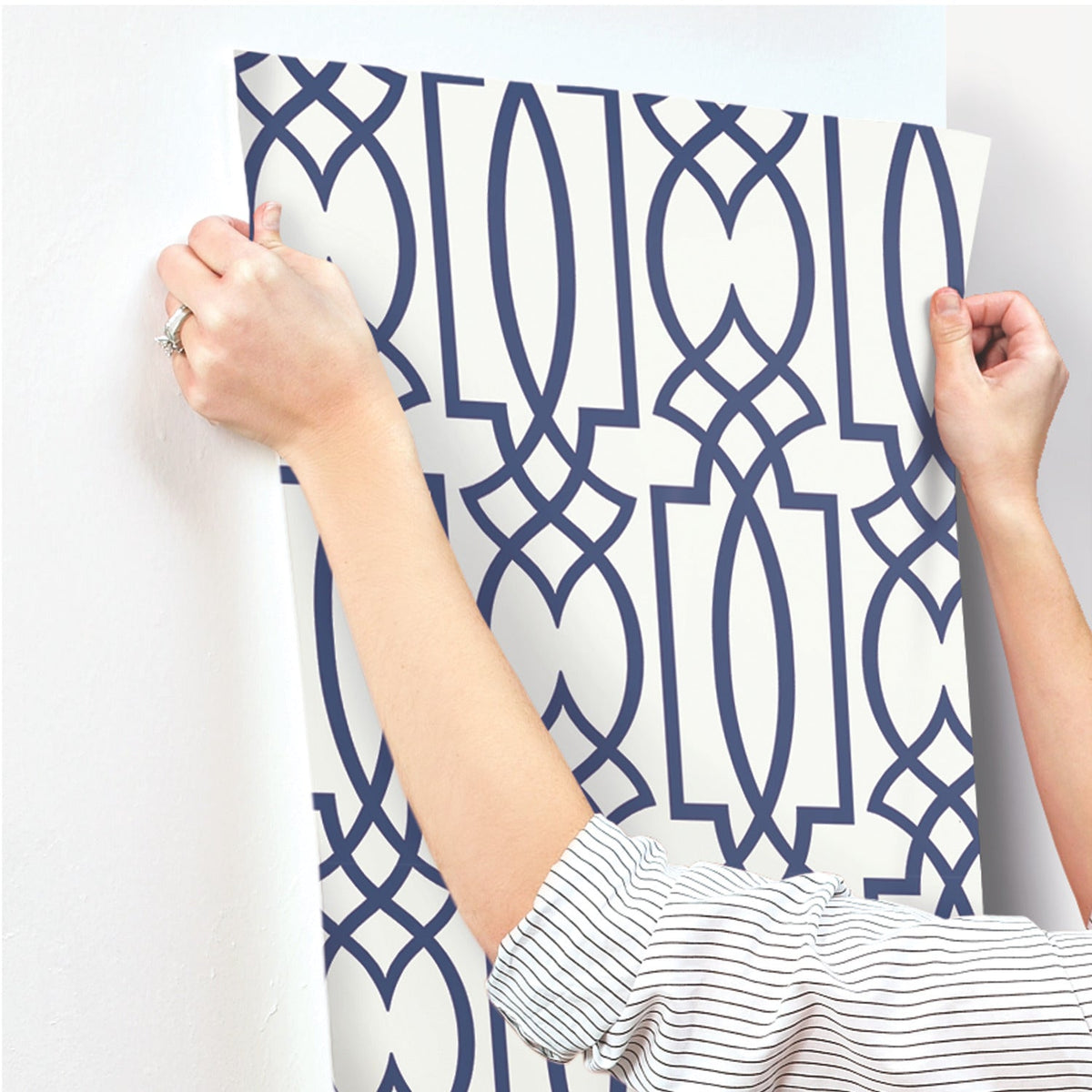 Hands applying a sheet of off-white Large Lattice Wallpaper with a bold blue geometric lattice pattern to a white wall.
