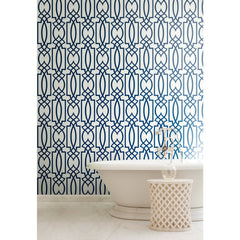 A white freestanding bathtub and small side table set against a dark blue Large Lattice Wallpaper by York Wallcoverings with a repeating geometric pattern.