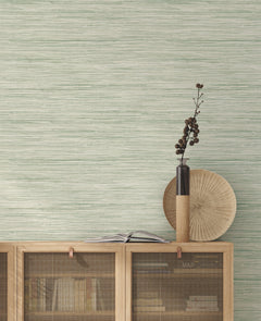 A minimalist interior featuring a light wood cabinet, books, an open book, a tall vase with dried branches, and a round wooden plate is set against a green textured wall adorned with York Wallcoverings’ Bahiagrass Wallpaper.