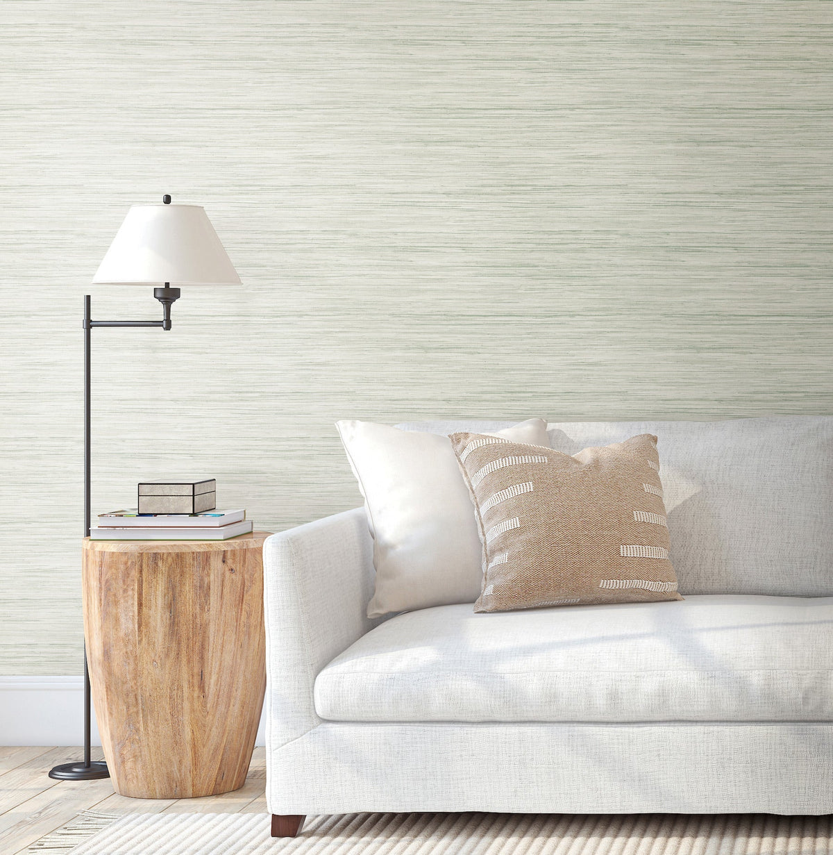 A light gray sofa with white and beige pillows sits next to a wooden side table with books and a black floor lamp, set against a wall adorned with Bahiagrass Wallpaper by York Wallcoverings.