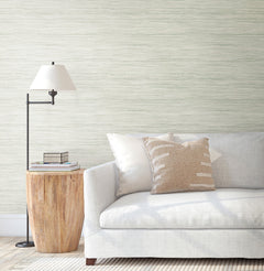 A modern living room features a white sofa, beige-patterned pillow, wooden side table with books, and a floor lamp, set against walls adorned with York Wallcoverings’ Bahiagrass Wallpaper and complemented by a neutral rug.