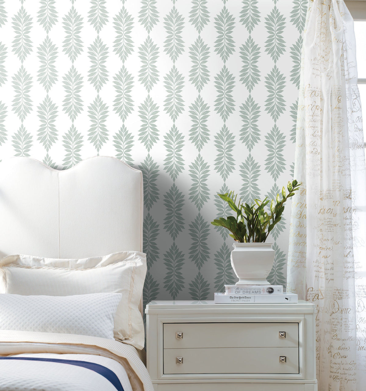 A white bed and nightstand sit next to a plant in a white pot. The wall is decorated with Broadsands Botanica Wallpaper by York Wallcoverings, featuring a repeating leafy pattern, while sheer curtains with script text hang on the right. The scene looks clean and bright.