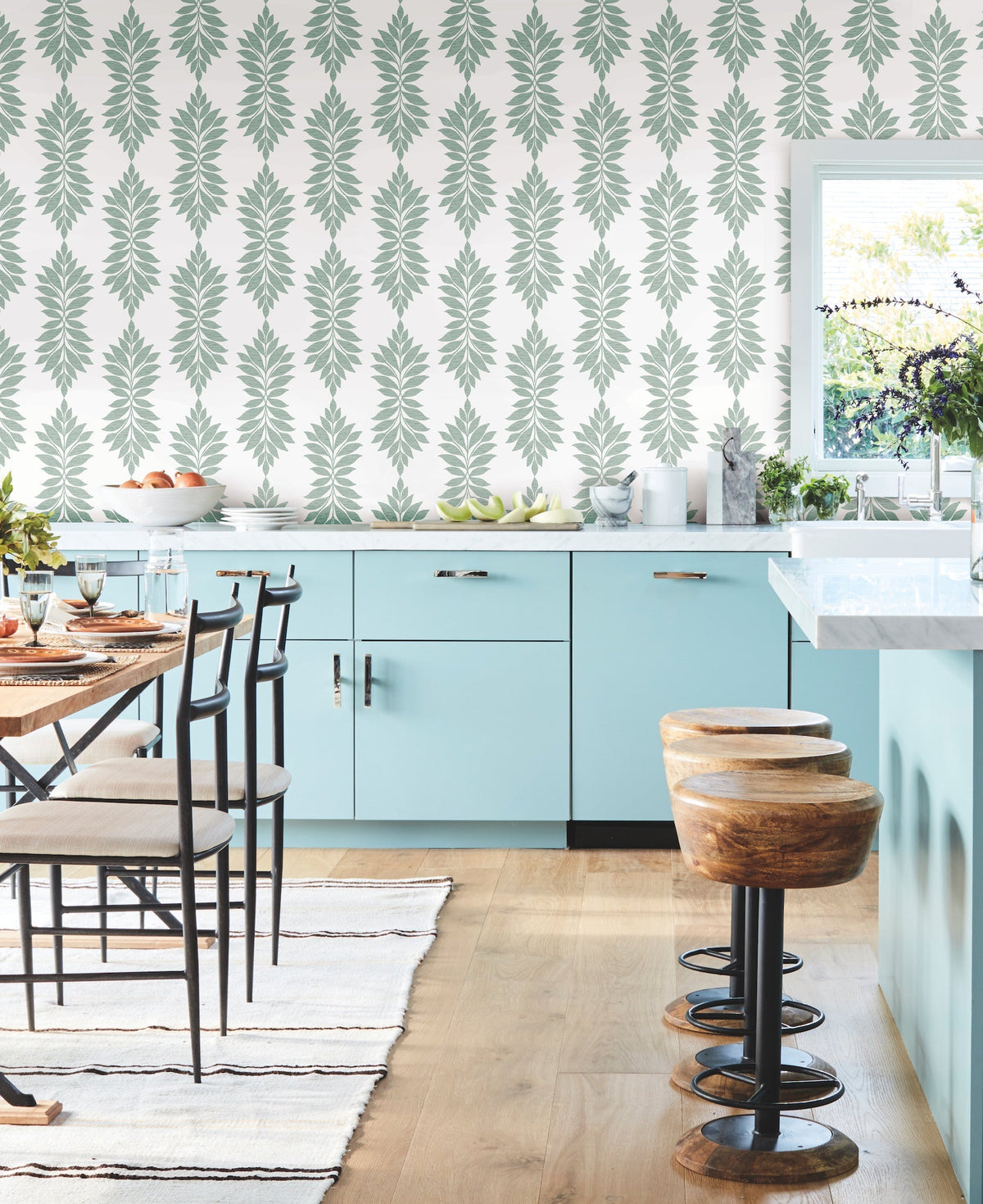 Modern kitchen with light blue cabinets, wooden stools, a white island, Broadsands Botanica Wallpaper by York Wallcoverings, a striped rug, and a window letting in natural light. A dining table set with plates and glasses is visible on the left side.