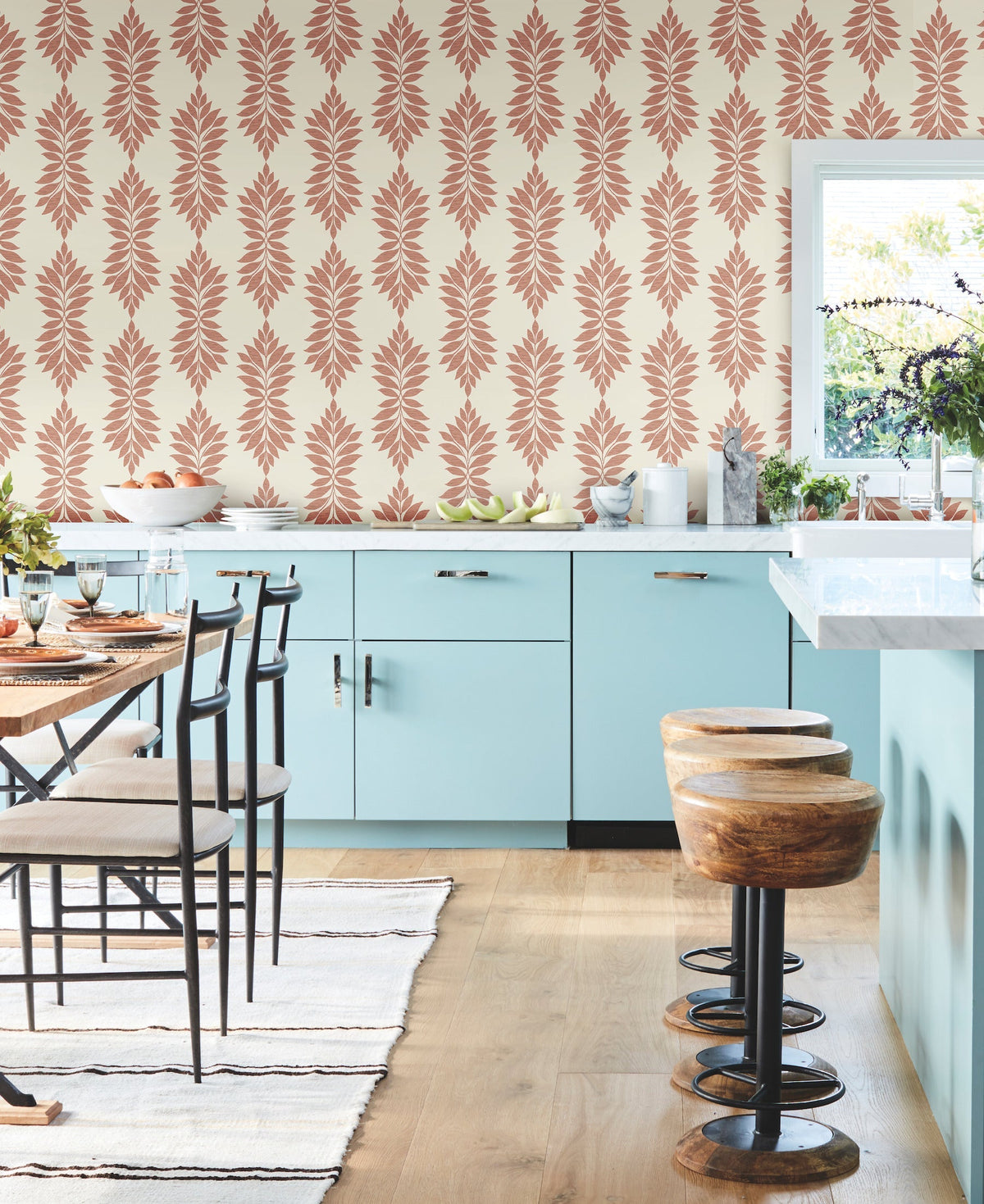 Modern kitchen with light blue cabinets, wooden stools, and a striped rug. The walls are adorned with Broadsands Botanica Wallpaper by York Wallcoverings in pink tones. A large window lets in natural light, and the dining table with chairs is set for a meal.