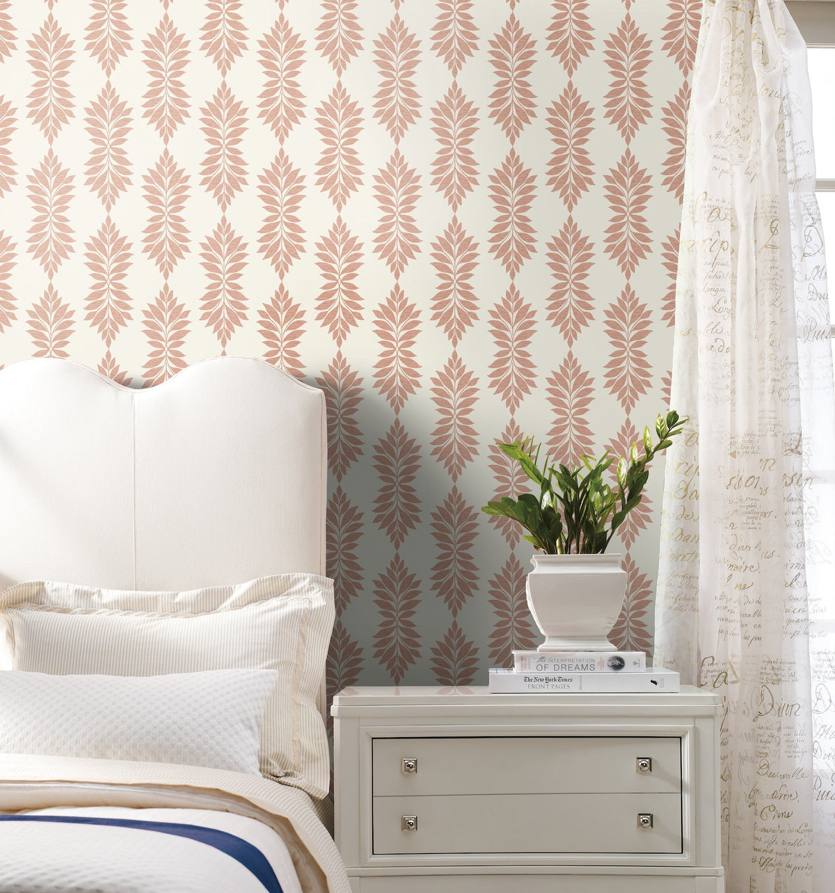 A white bed and nightstand are placed beside a potted plant in a bright bedroom, featuring Broadsands Botanica Wallpaper by York Wallcoverings, with cream and pink patterns, and sheer script curtains.