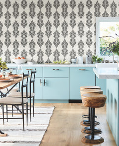 Modern kitchen with light blue cabinets, Broadsands Botanica Wallpaper by York Wallcoverings in gray and white, wooden stools, dining table with chairs, striped rug, and a window letting in natural light.
