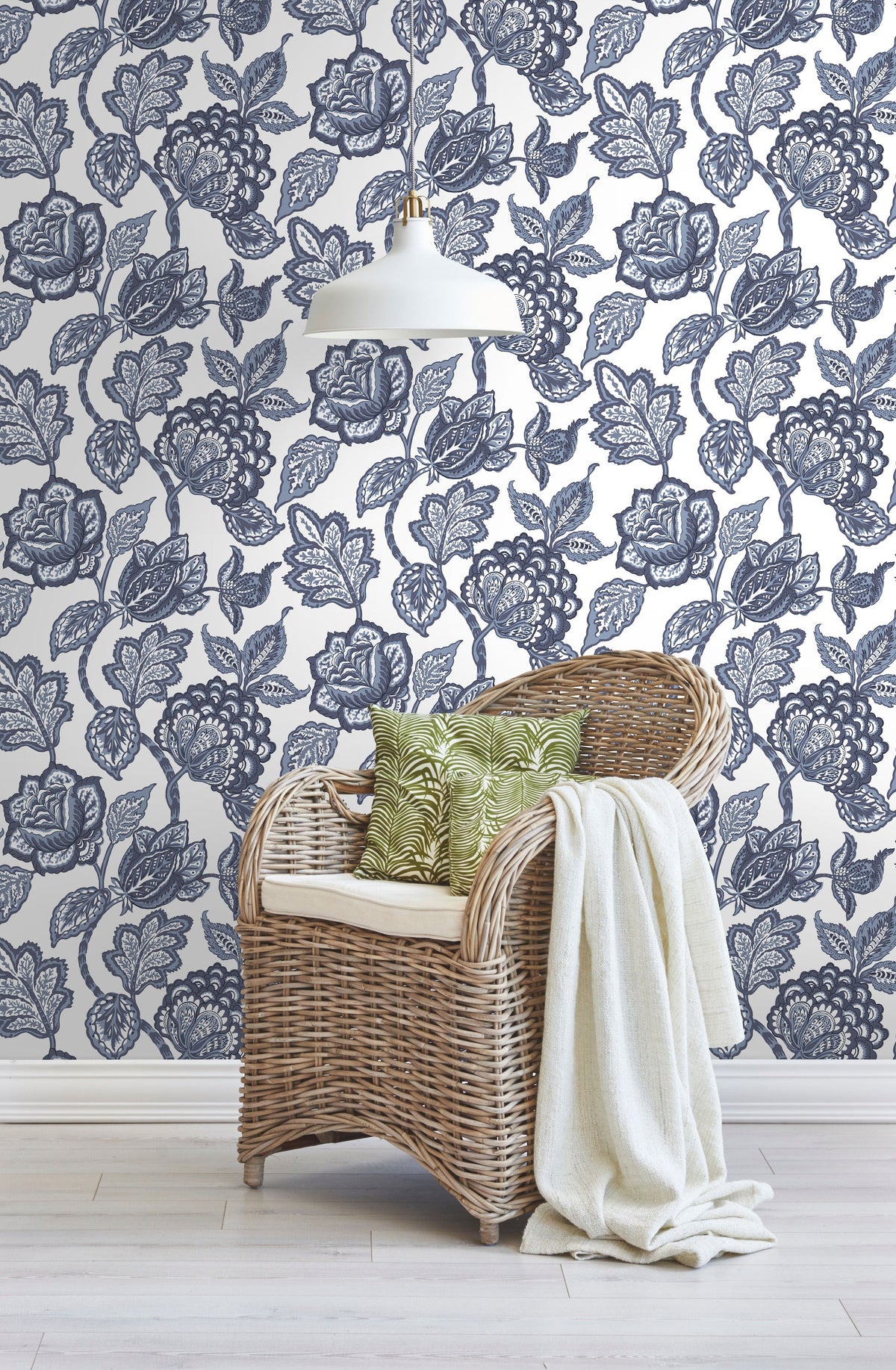 A wicker chair with a white cushion, green patterned pillows, and a cream throw blanket sits in front of Midsummer Jacobean Wallpaper by York Wallcoverings, featuring a dark blue botanical print of flowers and vines.